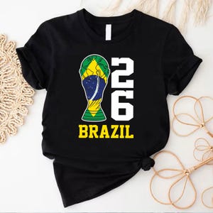 May include: A black short-sleeved t-shirt featuring a graphic of a trophy cup filled with the green, yellow, and blue colors of the Brazilian flag. To the right of the trophy is the number 26 in white, with the word BRAZIL printed in yellow below.