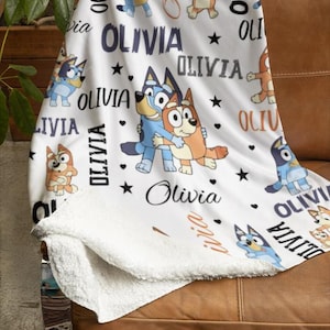 May include: White fleece blanket featuring cartoon characters and the name "Olivia" in various fonts. The blanket has a soft, plush texture and is decorated with stars and hearts. The blanket is folded over a brown leather couch.