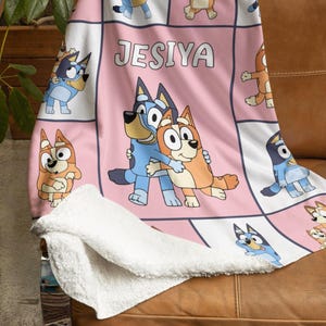 Könnte beinhalten: Eine rosa-weiße Decke mit Cartoon-Figuren und dem Namen "JESIYA". Die Decke hat eine weiche, weiße, plüschige Unterseite und ist über ein braunes Ledersofa drapiert. Die Decke ist mit Cartoon-Hundefiguren verziert.