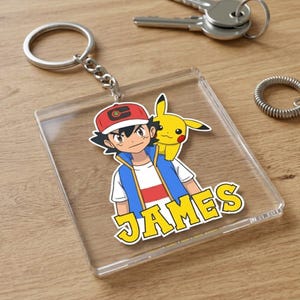 Puede incluir: Un llavero cuadrado de acrílico transparente con una ilustración colorida de un personaje de dibujos animados con gorra roja y una criatura amarilla sobre el hombro. El nombre JAMES está impreso en letras amarillas con borde blanco.
