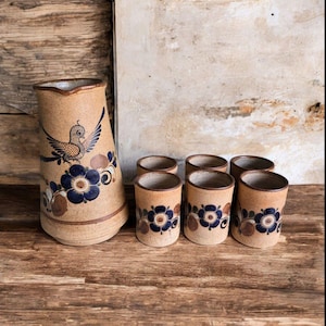 May include: A tan ceramic pitcher and six matching cups. The pitcher features a bird and floral design in blue and brown. The cups have a similar floral pattern. The set is displayed on a wooden surface.