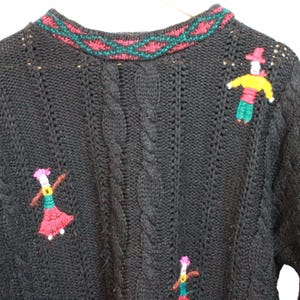 May include: Black knitted sweater with a cable knit pattern and embroidered figures. The neckline has red and green detailing. The sweater features several embroidered figures in various colours.