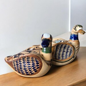 May include: Two ceramic ducks with hand-painted designs in shades of blue, brown, and white. The ducks are sitting on a wooden surface.