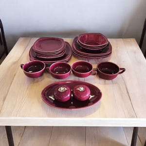 May include: A set of burgundy-colored ceramic dinnerware, including plates, bowls, and cups. The set is arranged on a wooden table.