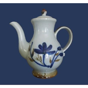 May include: White ceramic teapot with a blue floral design and brown accents. The teapot has a curved spout, a handle, and a lid with a brown knob. The base of the teapot is a light brown color.
