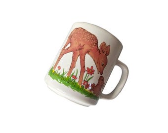 May include: White ceramic mug featuring a fawn and a small brown animal in a grassy scene with red flowers. The fawn is light brown with darker spots. The mug has a curved handle and a white background.