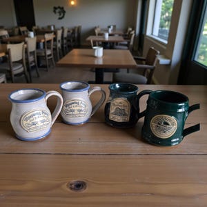 May include: Four ceramic mugs with handles are displayed on a wooden table. Two mugs are cream-coloured with blue rims and the text "Whispering Pines Bed & Breakfast." The other two are dark green with labels that read "The Rivertown Inn" and "Schumachers."
