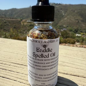 Könnte beinhalten: Eine klare Glasflasche mit schwarzem Tropfaufsatz, gefüllt mit hellfarbigem Öl und Pflanzen. Auf dem Etikett steht "Baddie Spelled Oil" und listet Inhaltsstoffe und Anweisungen auf.