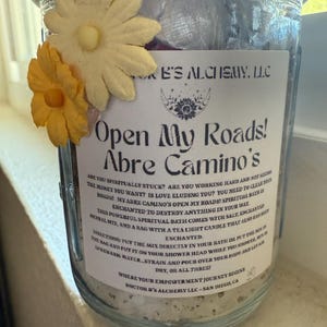 Pode incluir: Um frasco de vidro cheio de uma substância branca e granulada. O frasco tem um rótulo branco com o texto "Open My Roads! Abre Camino's" e instruções para um banho espiritual. O frasco é decorado com duas flores amarelas.