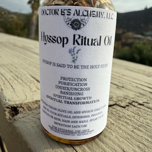 May include: A clear glass bottle with a white label that reads "Hyssop Ritual Oil". The label also includes the text "Doctor B's Alchemy, LLC" and "Hyssop is said to be the holy herb". The bottle contains a golden liquid and is sitting on a wooden surface.