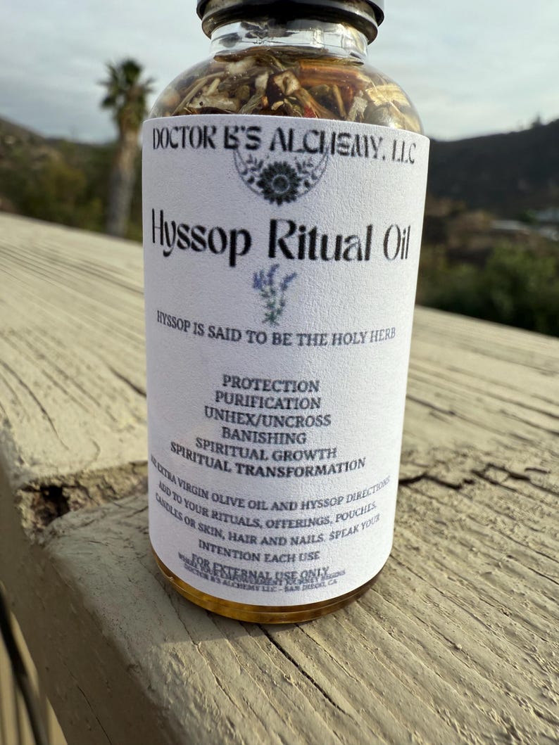 May include: A clear glass bottle with a white label that reads "Hyssop Ritual Oil". The label also includes the text "Doctor B's Alchemy, LLC" and "Hyssop is said to be the holy herb". The bottle contains a golden liquid and is sitting on a wooden surface.