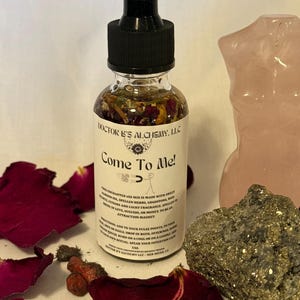 Op de afbeelding: Een heldere glazen fles met een zwarte druppelkap, gevuld met een vloeistof met kruiden en bloemblaadjes. Op het etiket staat "Come To Me!" en "Doctor B's Alchemy, LLC". Verspreid liggen rozenblaadjes, een roze stenen beeldhouwwerk en een pyrietsteen.