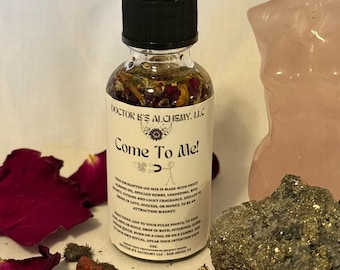 May include: A clear glass bottle with a black dropper top, filled with a liquid containing herbs and flower petals. The label reads "Come To Me!" and "Doctor B's Alchemy, LLC". Scattered around are rose petals, a pink stone carving, and a pyrite stone.