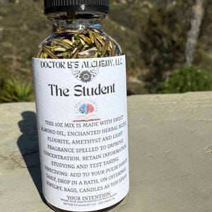 Puede incluir: Una pequeña botella de vidrio transparente con una tapa negra, etiquetada como "The Student" y "Doctor B's Alchemy, LLC". La botella contiene una mezcla de hierbas y aceites, y la etiqueta la describe como una mezcla de 30 ml hecha con aceite de almendras dulces, mezcla de hierbas encantadas, fluorita, amatista y fragancia ligera diseñada para mejorar la concentración, retener información, estudiar y realizar pruebas. La etiqueta también incluye instrucciones de uso, como agregar a los puntos de pulso diariamente, dejar caer en un baño o usar como ofrendas.
