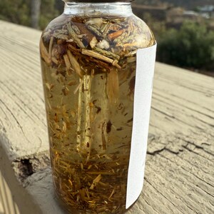 May include: A clear glass bottle with a black lid filled with a light brown liquid and various herbs and plant matter. The bottle is sitting on a wooden surface.