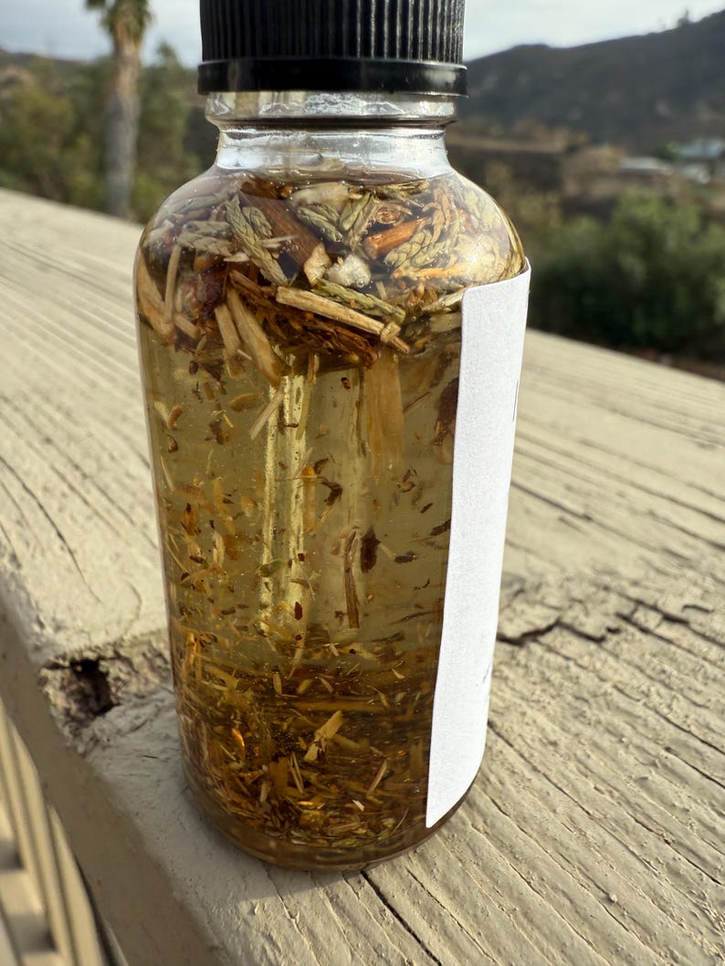 May include: A clear glass bottle with a black lid filled with a light brown liquid and various herbs and plant matter. The bottle is sitting on a wooden surface.