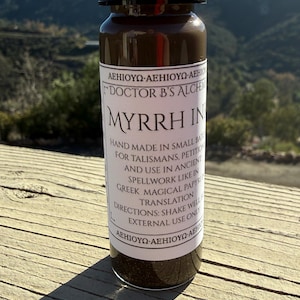 May include: A glass bottle of Myrrh Ink with a black cap, labeled "Doctor B's Alchemy." The label includes text about hand-made ink for talismans and spellwork. The bottle is on a wooden surface with a mountain backdrop.