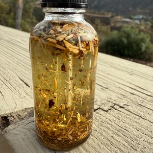May include: A clear glass bottle with a black lid filled with a golden liquid and various herbs and botanicals. The bottle is sitting on a wooden surface.