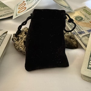 May include: A small, black velvet drawstring bag sits atop a cluster of pyrite stones, surrounded by several one hundred dollar bills. The bills are arranged on a white surface, creating a backdrop for the bag.