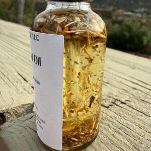 May include: A clear glass bottle with a black cap filled with a yellow liquid and herbs. The label on the bottle reads "Herb Oil" and "[Company Name] LLC".