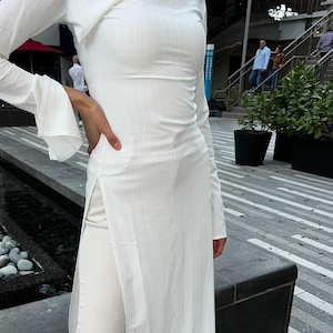 May include: A white long-sleeved tunic top with a side slit and a matching pair of white pants. The outfit is styled with white sneakers.