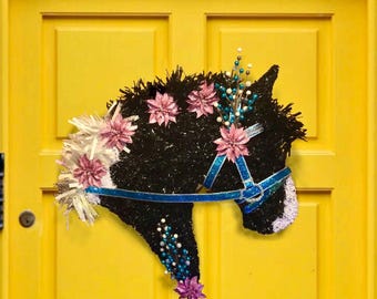 May include: A black horse head wreath with blue ribbon and pink flowers. The wreath is decorated with white and blue accents and is perfect for a front door.