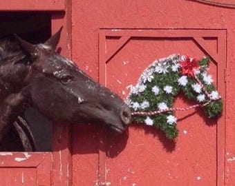 May include: A black horse's head peeks through a red wooden stall door. A green and white floral horse head wreath is attached to the door.