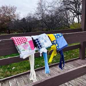 May include: Three colorful bonnets with ties hang on a wooden railing. The bonnets feature various patterns, including red gingham, floral prints, and patchwork designs. Each bonnet has long, fabric ties in shades of white, light blue, and yellow.