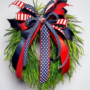 May include: A festive wreath with a base of green foliage, adorned with red, white, and blue ribbons. The ribbons feature star and stripe patterns, creating a patriotic theme. The wreath is suitable for seasonal or holiday decor.