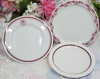 May include: Three white ceramic plates with a red rim. The center plate has a red design with a stylized flower pattern. The plate on the left has a red rim and a red logo with the letters "U.S." and a stylized caduceus symbol. The plate on the right has a red rim.