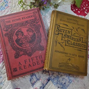 Pode incluir: Dois livros antigos de capa dura, um vermelho com letras douradas intitulado "The Howe Readers A Fifth Reader" e o outro dourado com letras pretas intitulado "Standard Supplementary Readers Seven American Classics Supplementary to Fifth Reader".