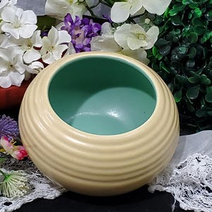 May include: A round, cream-colored ceramic bowl with a light green interior. The exterior features horizontal ridged detailing. The bowl is surrounded by white and purple flowers, green leaves, and a green topiary ball.