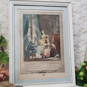 May include: A framed print titled LA TOILETTE featuring an 18th-century interior scene with three figures in period clothing. The artwork is encased in a white frame with a light blue mat, depicting a person seated at a vanity with an attendant.