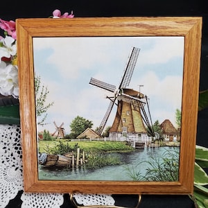 May include: A framed tile with a painting of a Dutch windmill in a rural landscape. The windmill is brown with white accents and has a blue sky and green grass in the background.