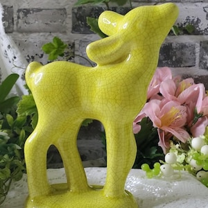 May include: A bright yellow ceramic deer figurine featuring a distinct crackle glaze finish. The deer is depicted in a standing position with its head tilted upward, showcasing a smooth, glossy surface texture and a solid, oval-shaped base.