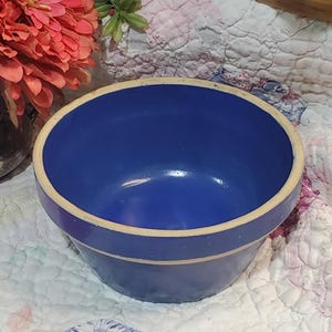 May include: A blue ceramic bowl with a cream-colored rim. The bowl has a glossy finish and a rounded shape. The interior is a deep blue, contrasting with the lighter rim. This vintage bowl is suitable for serving or display.