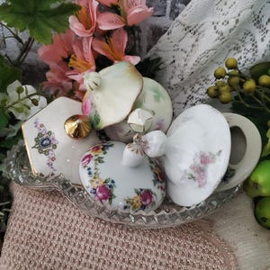 May include: A collection of four assorted porcelain lids with floral patterns and gold-tone accents, resting inside a clear, pressed glass dish with a scalloped edge. The lids feature various shapes, including round and hexagonal, with decorative knobs.