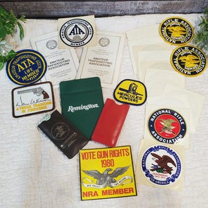 May include: A collection of vintage patches, stickers, and documents related to shooting sports. Items include patches for the Amateur Trapshooting Association, National Rifle Association, and Hercules Powders. Also included are a green Remington case and a red leather case.