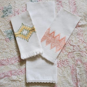 Op de afbeelding: Drie witte handdoeken met geborduurde patronen. Eén handdoek heeft een geel en groen geometrisch ontwerp, een andere een koraal zigzagpatroon, en de derde is effen. De handdoeken hebben gefranjerde randen en decoratieve afwerking.