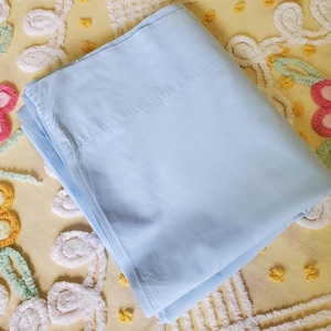 May include: A folded light blue fabric sheet with a smooth, matte texture. The textile is rectangular in shape, featuring clean, straight edges and a consistent solid color throughout, showing a simple and functional design for bedding or home use.