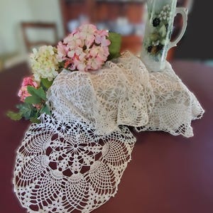 May include: A vintage-style display featuring a white crocheted lace doily and a matching draped cloth. A decorative pitcher with floral and berry designs sits atop the cloth, alongside pink and white hydrangeas. The arrangement is set on a dark red surface.