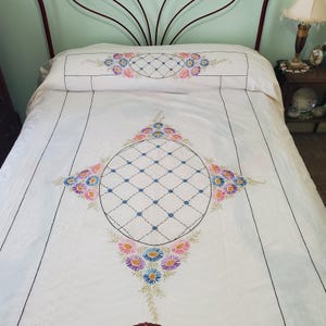 Op de afbeelding: Wit sprei met geborduurde bloemen- en geometrische patronen. Het sprei heeft een grote centrale ovaal met een ruitpatroon, omringd door kleurrijke bloemen in roze, blauw en paars. Zwarte lijnen omlijsten het ontwerp.