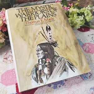 Może przedstawiać: Twarda oprawa książki zatytułowanej "The Mystic Warriors of the Plains" autorstwa Thomasa E. Mailsa. Okładka przedstawia ilustrację Indianina w profilu, ozdobionego piórami. Tytuł książki jest dużymi, pogrubionymi literami.