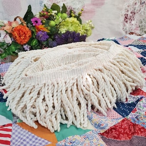 Op de afbeelding: Crèmekleurige, geweven franjeband met dikke, gedraaide, touwachtige strengen. De band is in een cirkelvormige vorm gerangschikt, met een geweven basis. De achtergrond omvat een kleurrijke quilt en een bloemenmand.