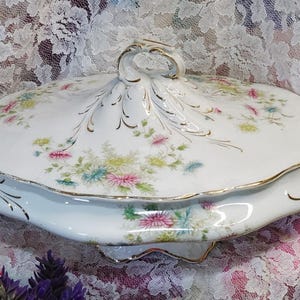 May include: A white porcelain serving dish with a lid, featuring a floral pattern in pink, blue, and yellow. The dish has gold trim and handles, and a decorative handle on the lid. The overall shape is oval, and the dish is likely vintage.