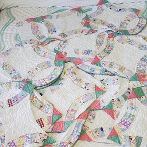 Pode incluir: Uma coleção de colchas feitas à mão com um padrão Double Wedding Ring. As colchas apresentam tecido branco com anéis de patchwork coloridos, triângulos rosa e verde e detalhes de acolchoado intrincados. As colchas são dispostas em uma colcha branca.