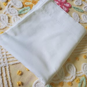 May include: A folded, solid white rectangular fabric sheet with a smooth, opaque texture. The fabric is neatly layered, showing a clean, straight hemmed edge along the top fold, presenting a simple and uniform appearance without any patterns or prints.