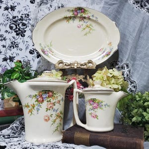 May include: A vintage cream-colored tea set with floral patterns. The set includes a large oval platter, a tall pitcher, and a smaller creamer. The floral design features pink, blue, and yellow flowers with gold trim.