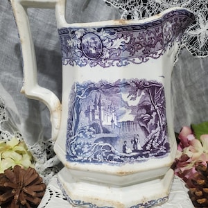 May include: Antique white and blue pitcher with a detailed landscape scene. The pitcher features a decorative handle and a wide spout. The landscape scene depicts a castle, trees, and figures. The pitcher has a decorative border.