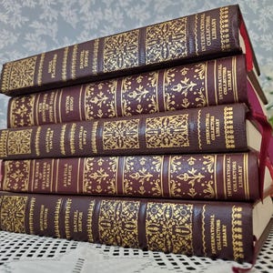 May include: A stack of five vintage books with burgundy covers and gold embossed floral designs. The spines feature titles and author names, including "Moll Flanders" and "Robinson Crusoe." Each book has a red ribbon bookmark and is part of the International Collectors Library.